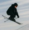 A freestyle skier mid-air above a snow ramp, frozen in perfect balance. The background is a blur of white and blue