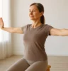 A wellness-focused adult performing posture exercises in a natural-light studio—seated or standing tall, with arms extended in a scapular squeeze, soft background