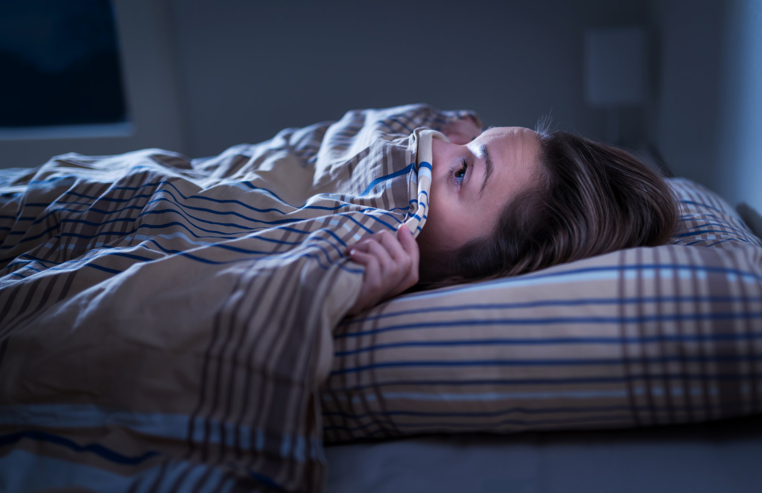 Woman in bed with duvet up over her nose, looking frightened.