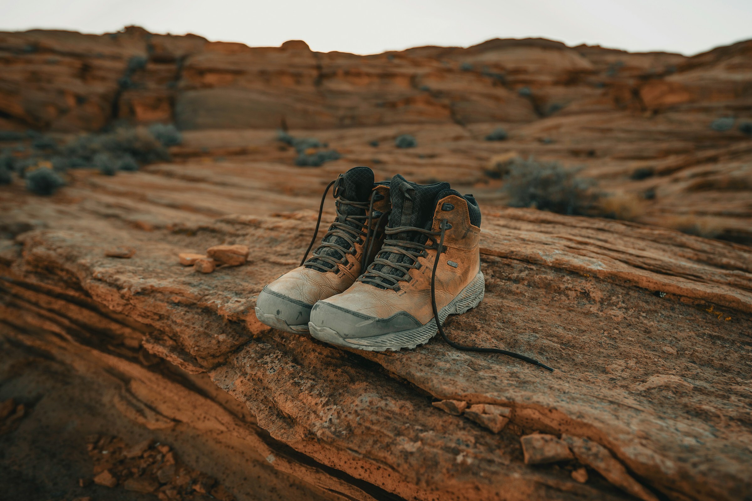 A pair of tan hiking boots with grey soles resting on layered red sandstone, with rocky desert terrain stretching behind them in soft light.