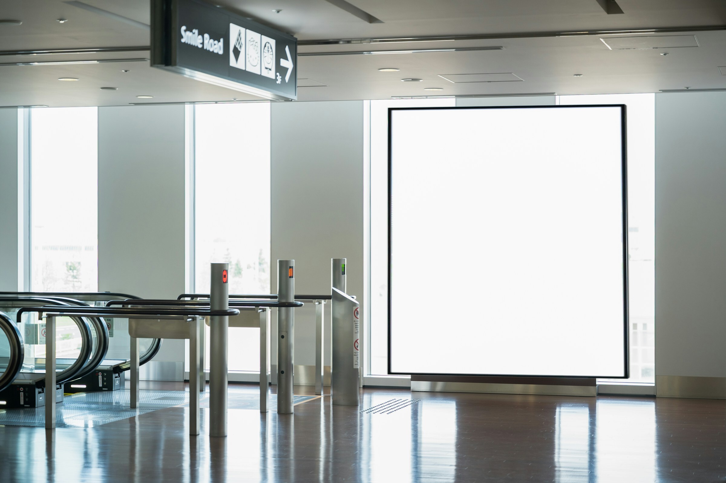 A large blank illuminated panel in an empty airport terminal beside escalators, with daylight through tall windows reflecting on the polished floor