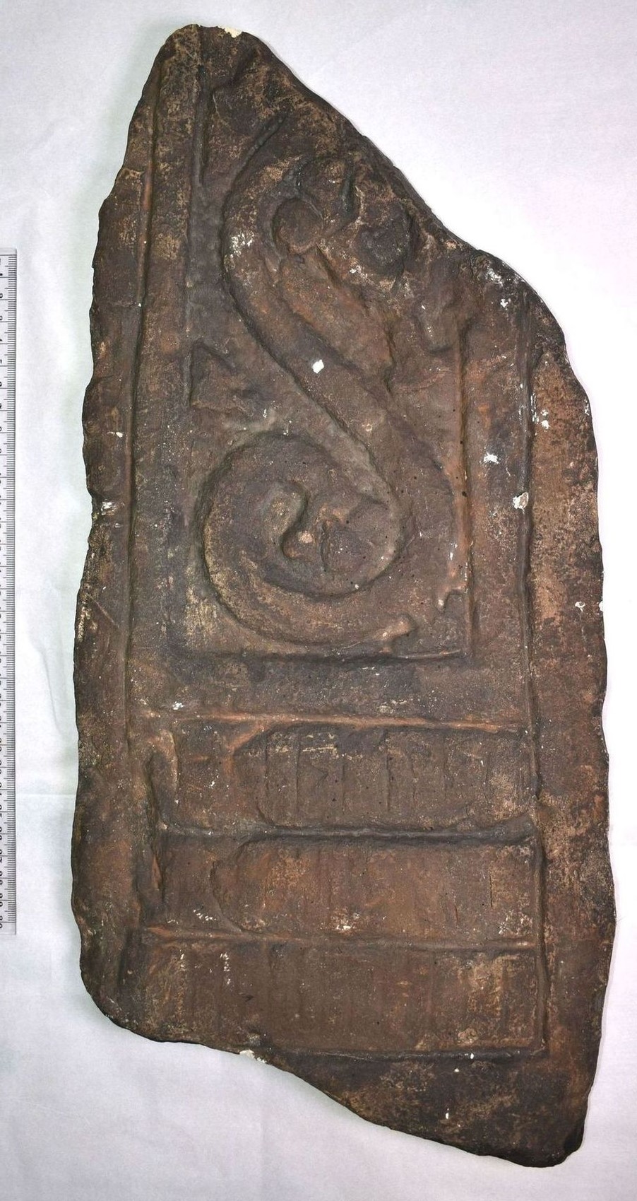 Ancient stone cast of part of a cross with intricate carvings, featuring a large spiral design and an inscription. It has a weathered texture and earthy tones, and a ruler for scale is partially visible on the left.