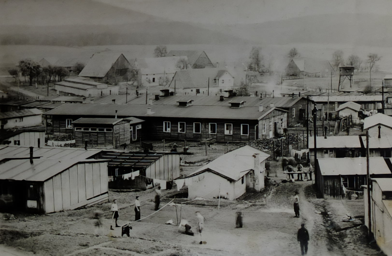 High view across a prisoner of war camp with rows of wooden huts, fences, a watchtower and farmland in the background.