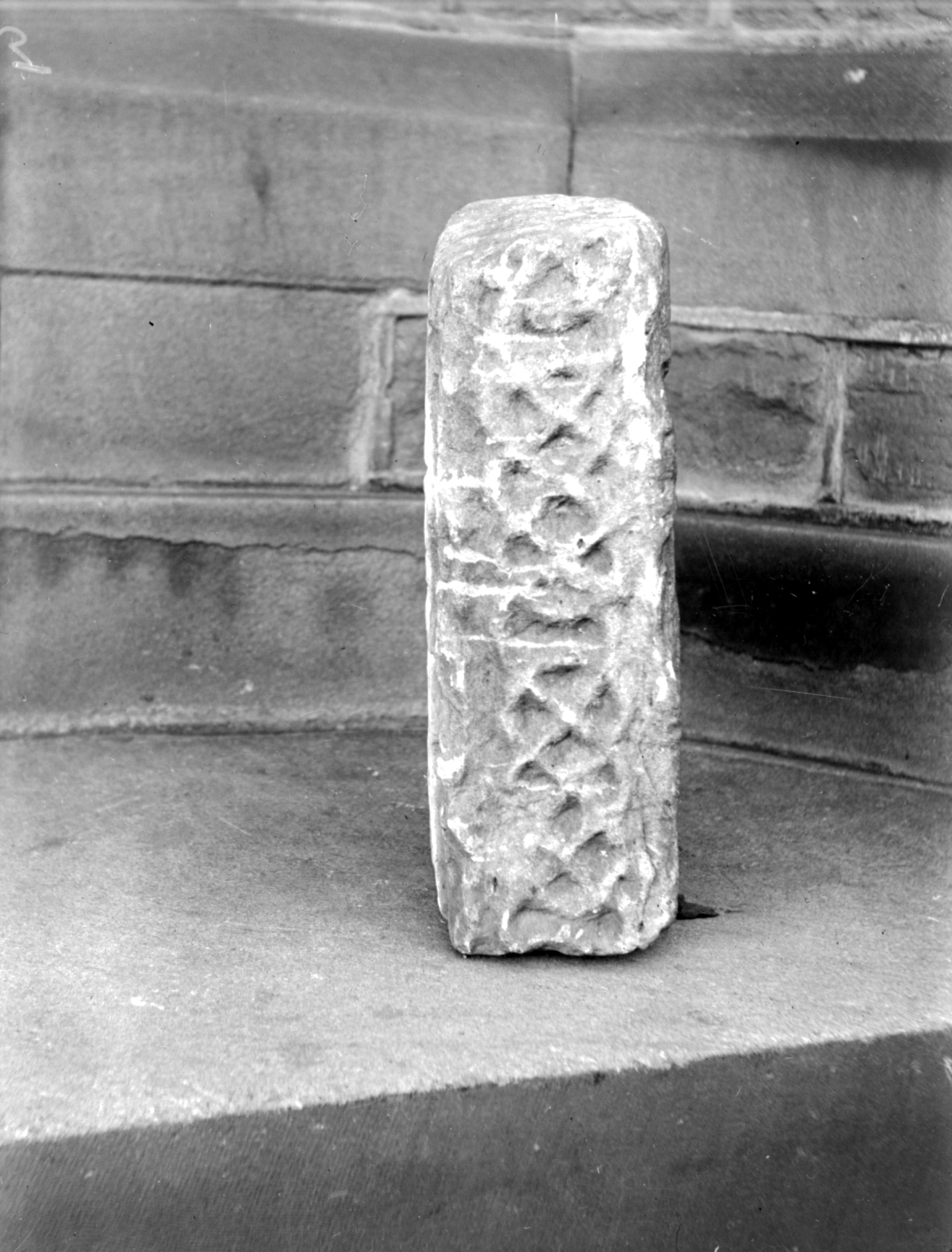 Black and white photograph of a Pre-Norman carved stone standing upright on a set of stone steps, featuring a worn interlaced pattern on its visible face. The background shows a stone wall with rough masonry and shallow relief detailing.
