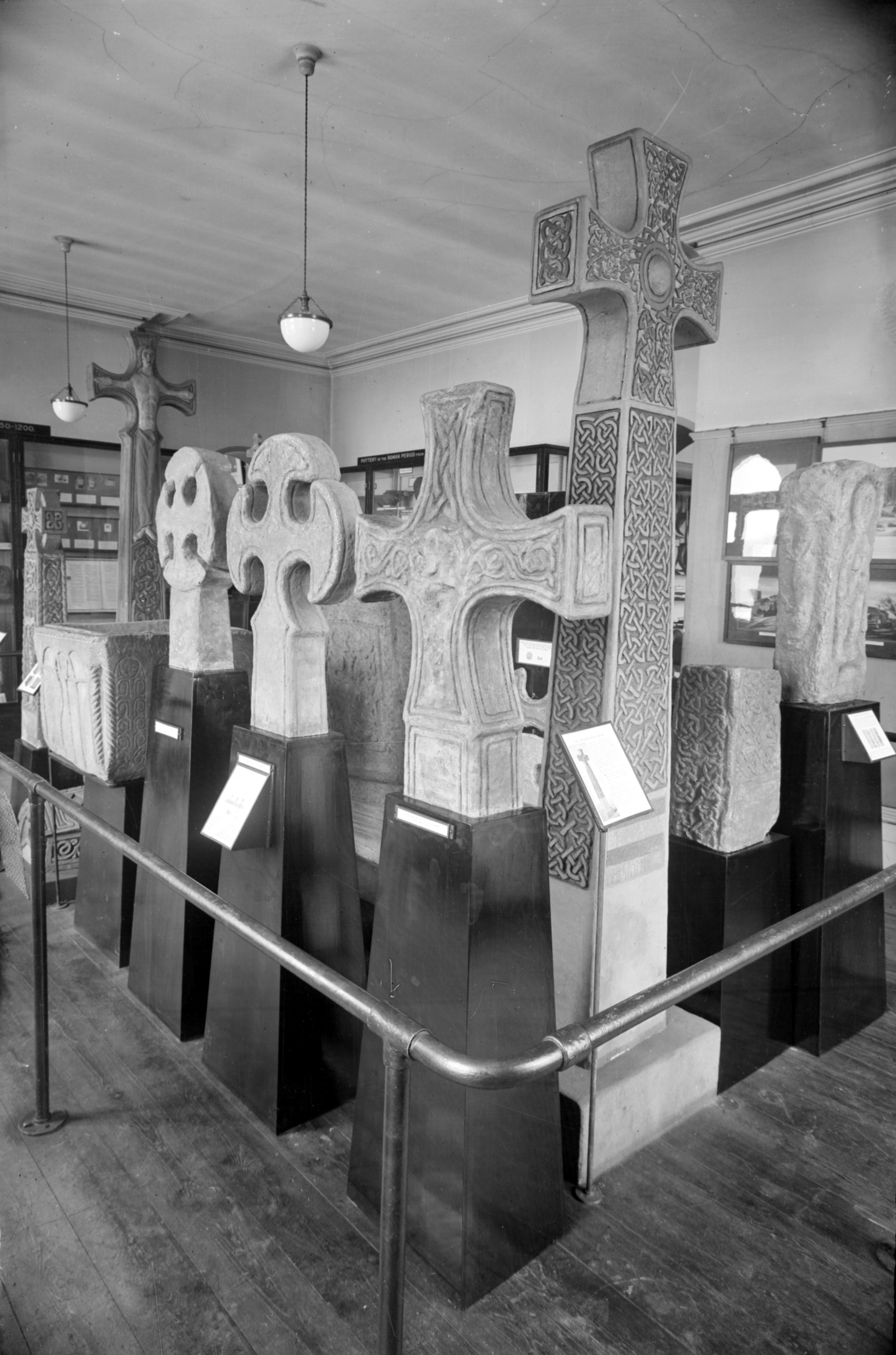 A black and white photograph of the Anglo-Saxon Room at Tolson Museum in Huddersfield circa 1931, displaying a row of intricately carved stone crosses and slabs on black pedestals behind a protective railing. The room features wooden floors, hanging light fixtures, and glass display cases lining the walls.