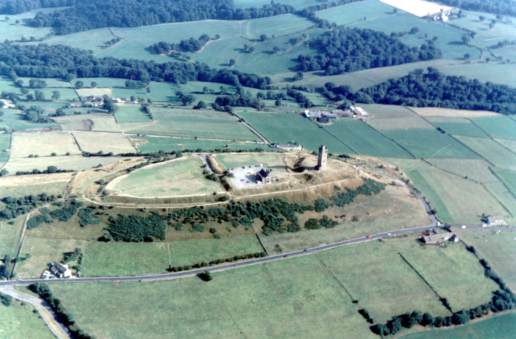 Aerial view of green countryside with rolling hills and scattered trees. A historic tower and ruins sit atop a hill, surrounded by fields and a road.