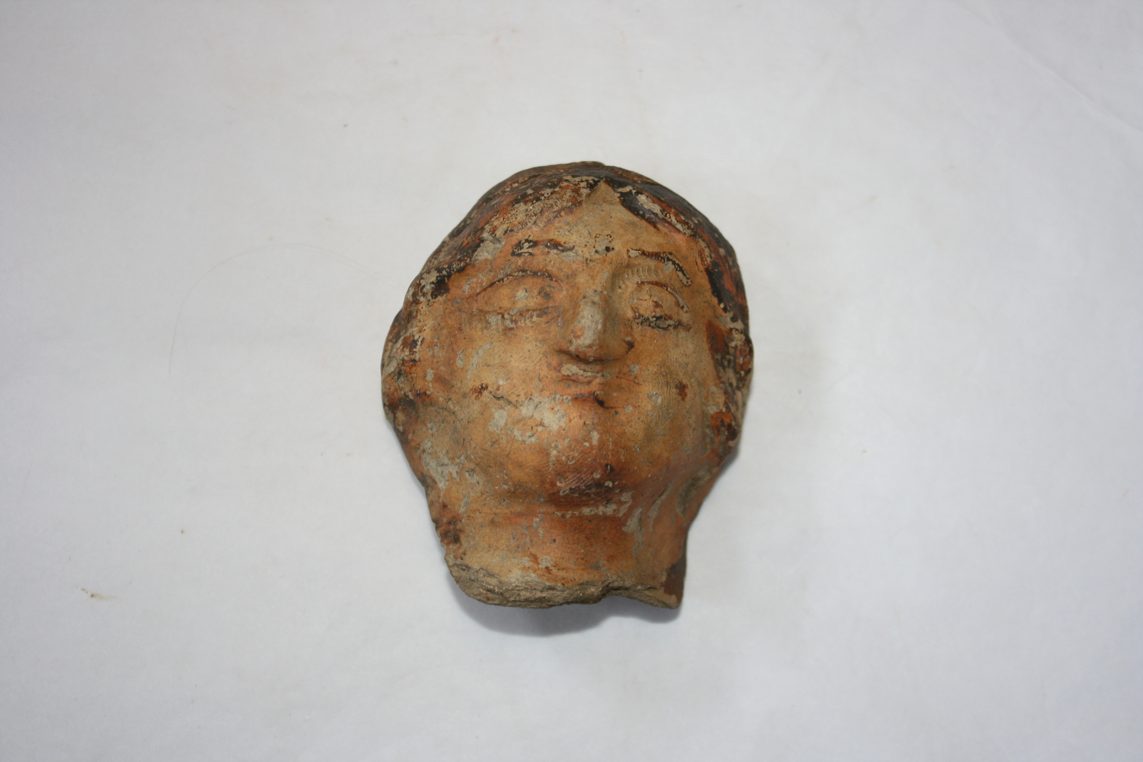 A colour photograph of a Roman ceramic head fragment placed on a light grey background. The terracotta head depicts a human face with faintly sculpted features and traces of paint on the surface.