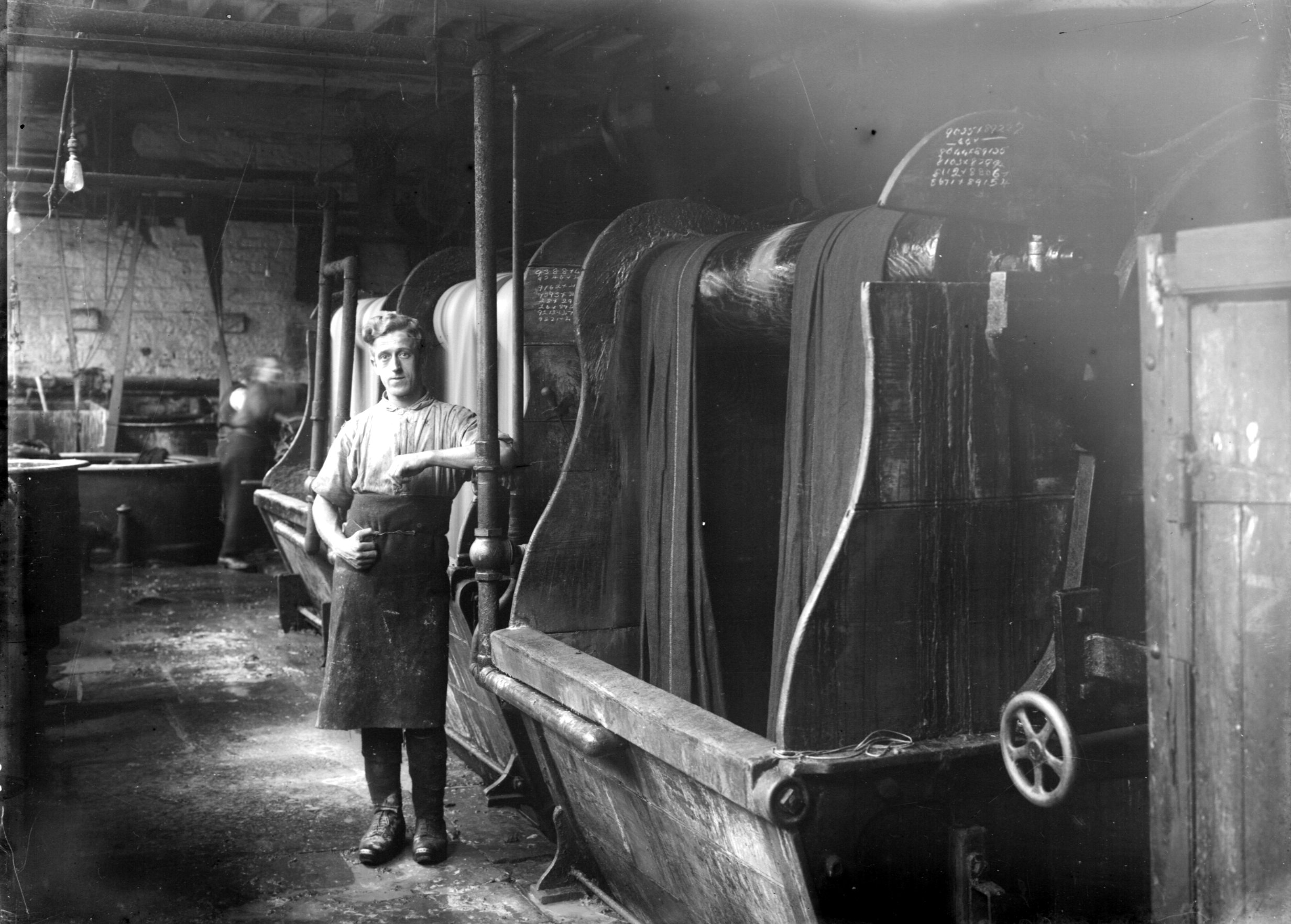 A black and white historical photograph showing a textile worker standing beside a large dyeing machine in a factory. The worker, wearing an apron and boots, poses with one hand on the machine. Long rolls of fabric are draped over the rotating drum of the dyeing equipment. The industrial setting is dimly lit, with visible pipes, brick walls, and other machinery in the background. Another worker appears blurred in motion further back, indicating activity in the workspace.