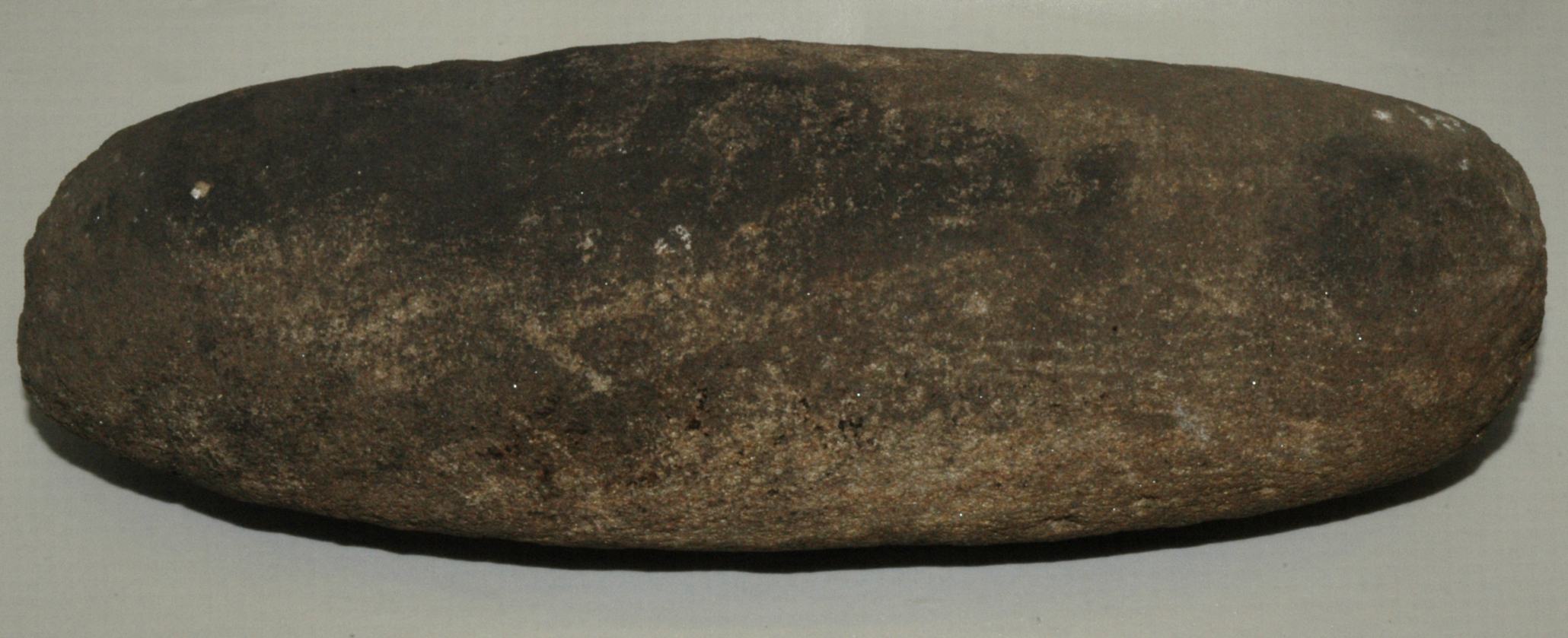 A long, cylindrical stone grain roller with rounded ends and a rough, textured surface showing signs of wear and age. The roller is dark brown with patches of lighter brown and some small white specks scattered across it, indicating its use over time. It lies horizontally against a plain light grey background.