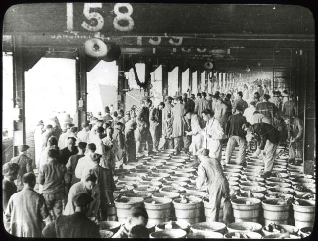 Black and white photograph showing an open warehouse with crowded with barrels of fish and men.