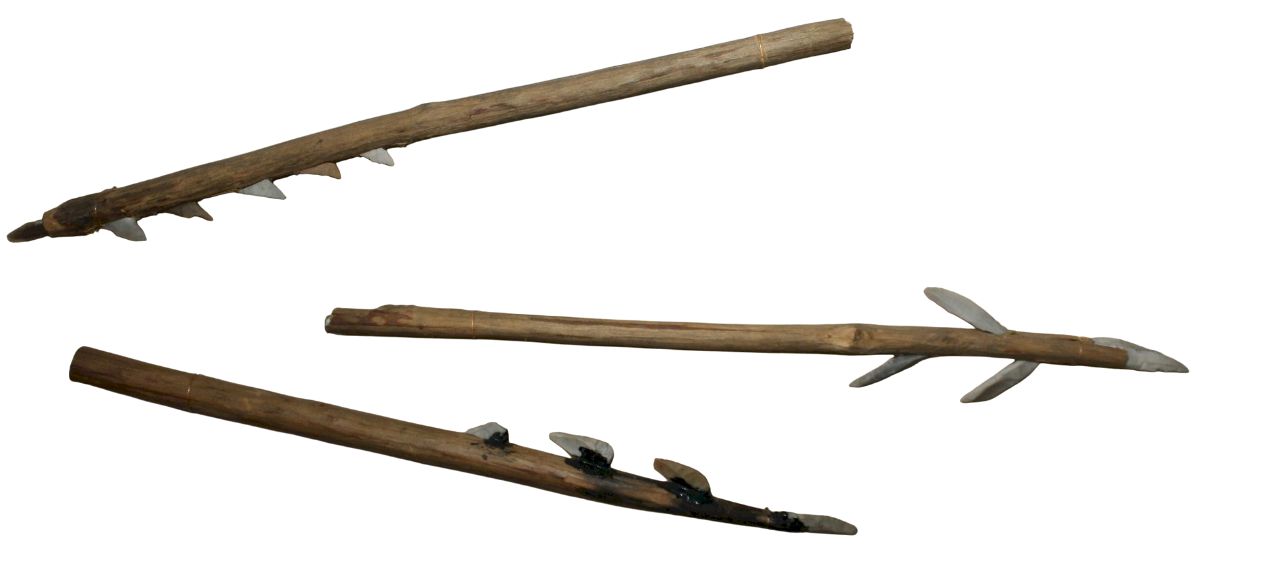 Three wooden fishing spears with multiple sharp, barbed flint points attached along the shafts, arranged horizontally on a white background. The spears vary slightly in length and design, with the flint points secured by bindings or resin, showing primitive craftsmanship typical of early fishing tools.
