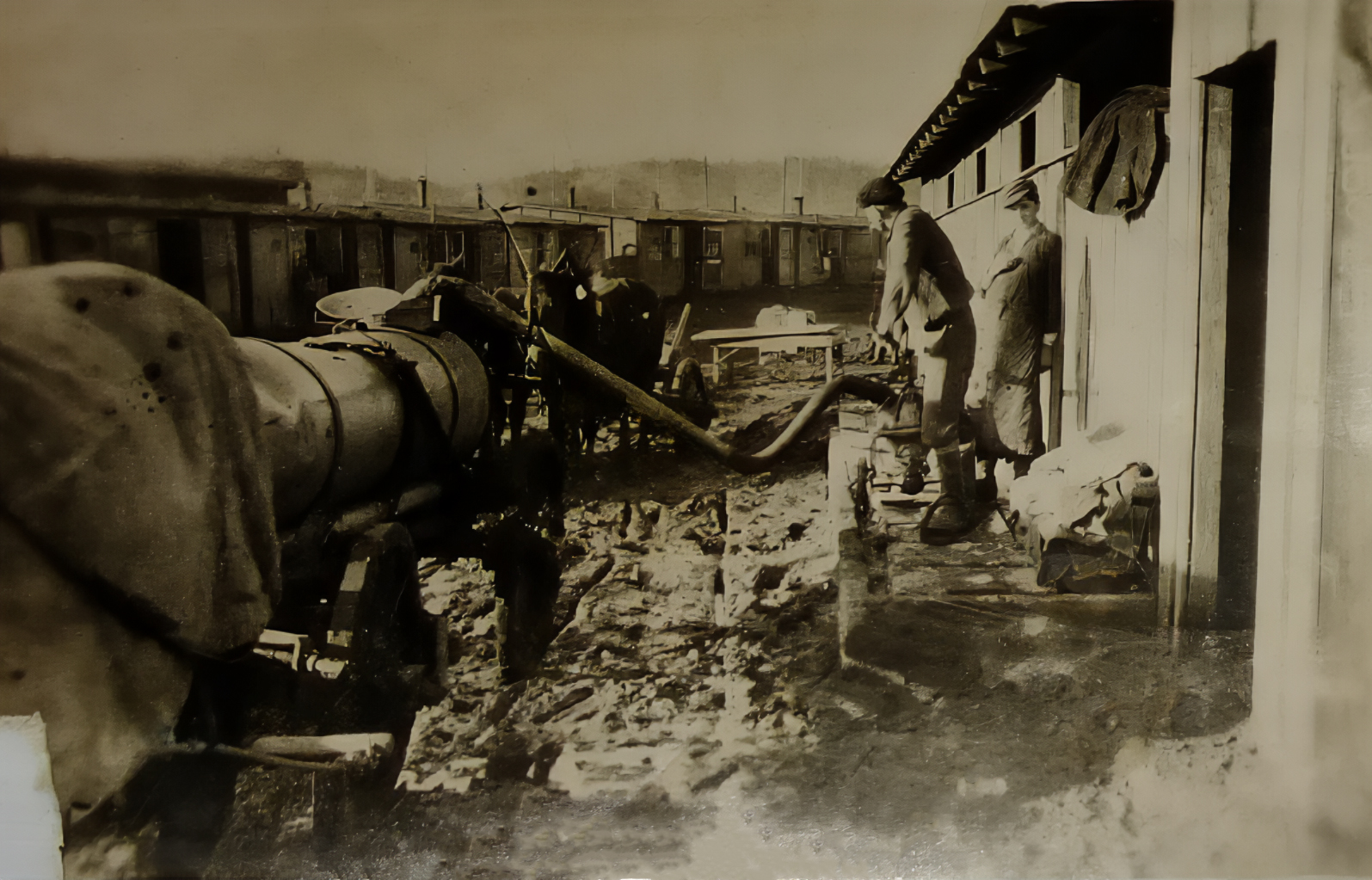 Prisoners working in a muddy yard between rows of wooden huts, using equipment beside a large pipe or boiler.