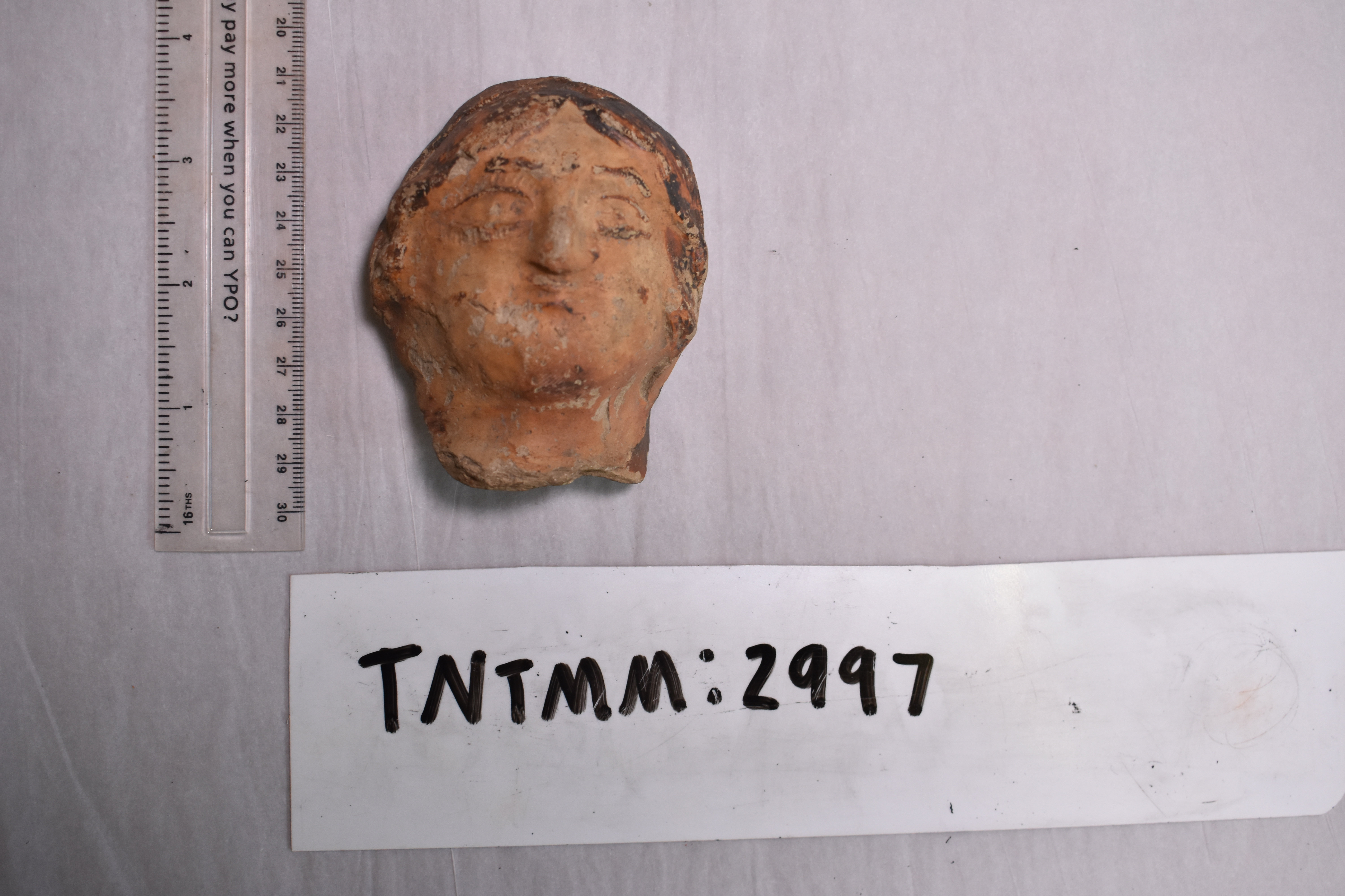 A colour photograph of a Roman ceramic head fragment placed on a light grey background. The terracotta head depicts a human face with faintly sculpted features and traces of paint on the surface. To the left of the object is a transparent plastic ruler marked in centimetres and inches for scale; the head is about 9 centimetres long. The head appears weathered and broken at the neck, indicating it was once part of a larger figure or statue. Below the artefact is a white label with the handwritten accession number for museum cataloguing.