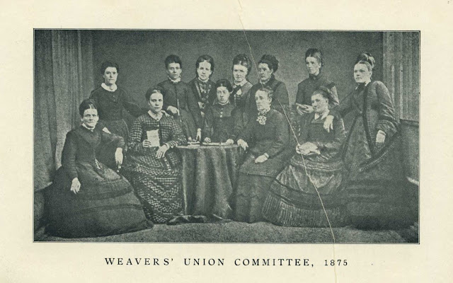 Black and white photograph of thirteen women in Victorian dress, seated and standing around a small table covered with a cloth. The women are formally posed, with neutral or serious expressions. The backdrop consists of heavy curtains. Printed text below the image reads in capital letters 'Weavers' Union Committee, 1875'.