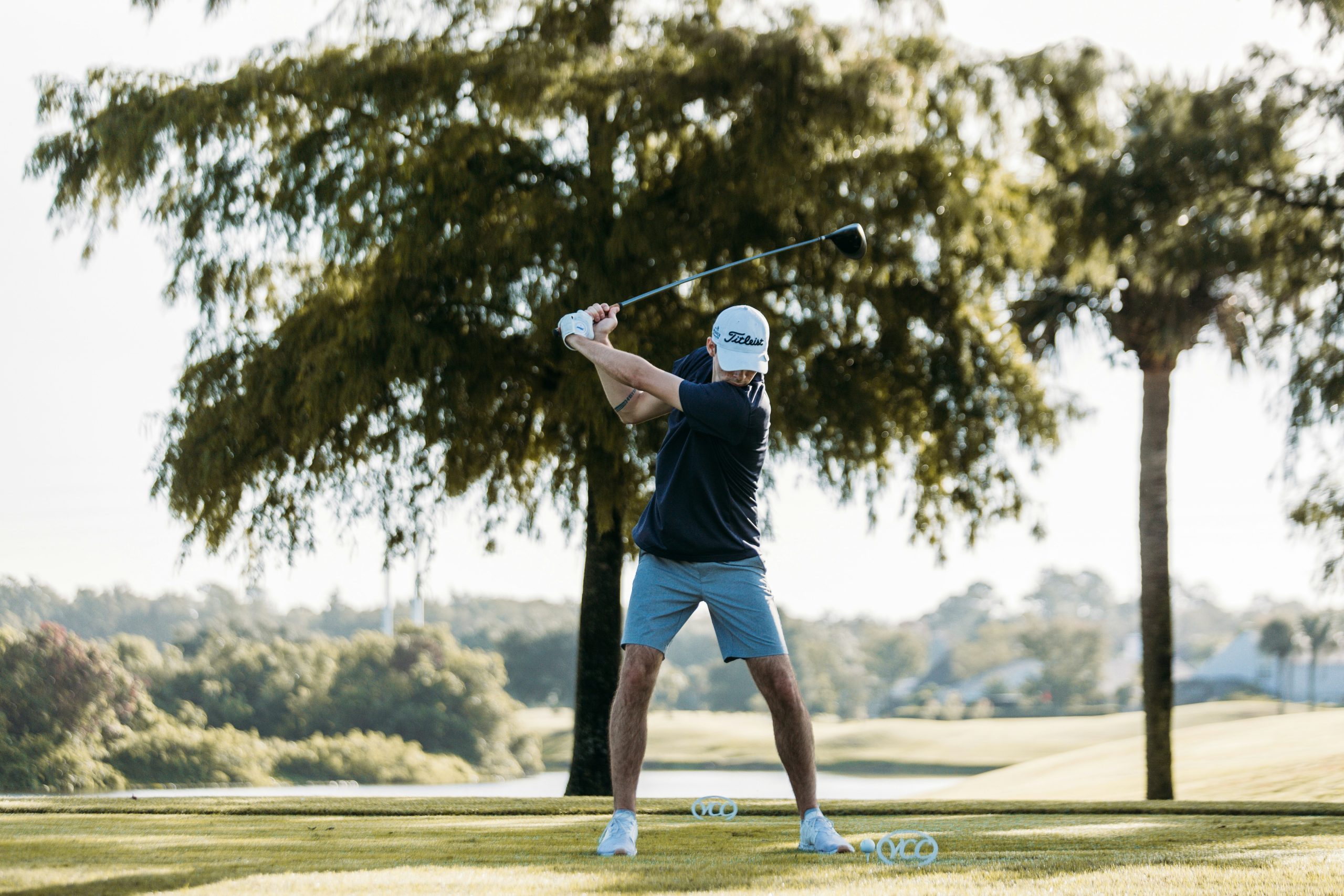 Golfer in mid-swing on a sunny golf course surrounded by trees and open landscape.