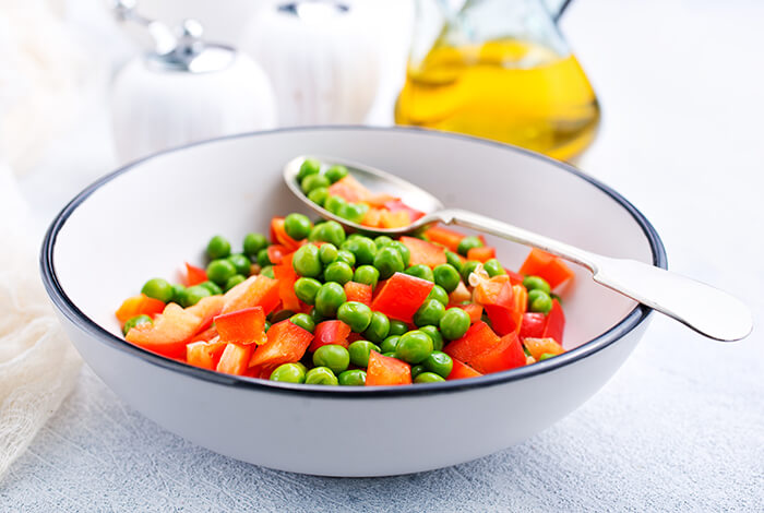 a bowl of green peas and chopped red bell pepper with a metal spoon