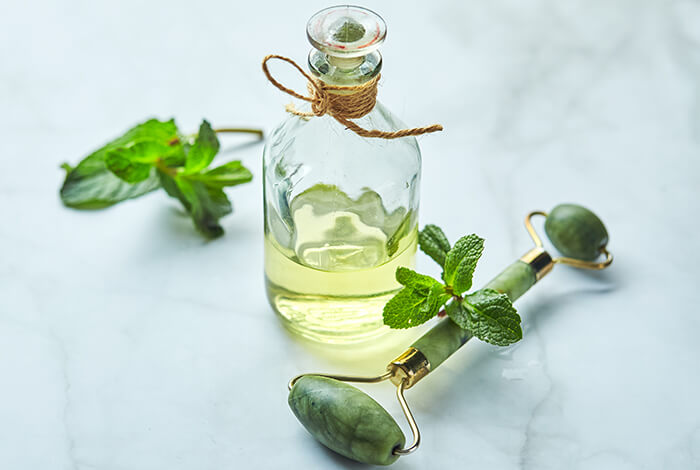 a bottle of mint essential oil alongside fresh green mint leaves and a jade face massage roller