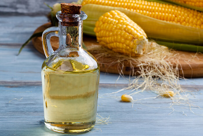 a bottle of corn oil placed in front of fresh corn cobs on a wooden surface