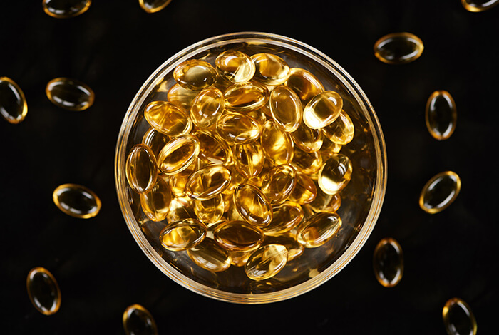 close-up of a glass bowl filled with golden fish oil capsules, with scattered capsules on a black background