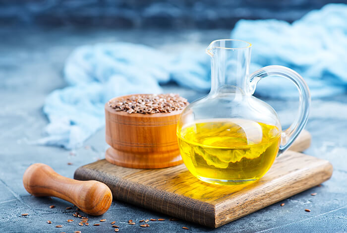 a glass pitcher of flaxseed oil on a wooden cutting board next to a round wooden container filled with flaxseeds