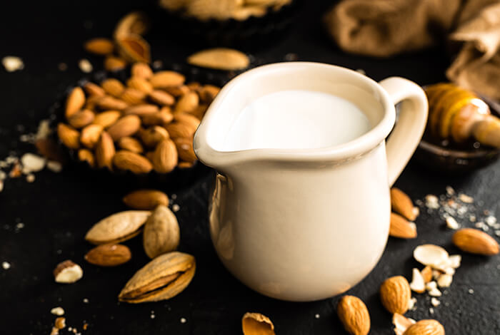 a white ceramic pitcher of almond milk, surrounded by almonds and a honey dipper on a dark background