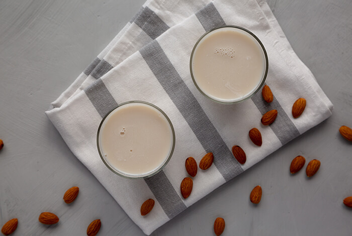 two glasses of almond milk on a striped dish towel, with scattered almonds on a gray background
