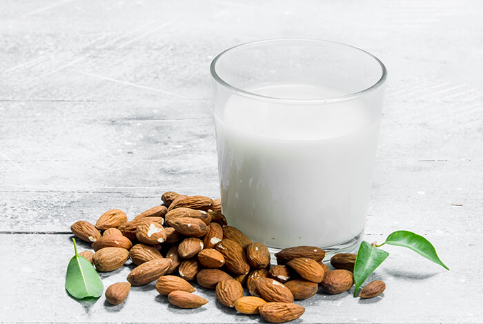 a glass of almond milk with raw almonds and green leaves on a light textured surface