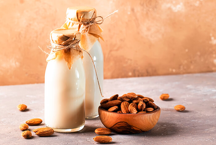 two glass bottles of almond milk with paper-tied tops next to a wooden bowl of almonds on a beige background
