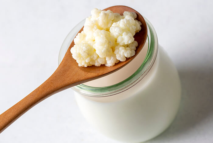 a wooden spoon holds white kefir grains above a clear glass jar filled with creamy, white kefir
