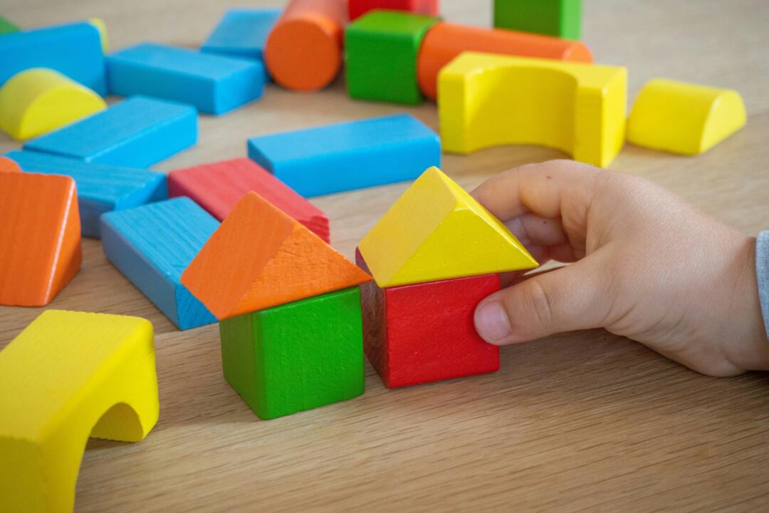 A child’s hand placing a yellow wooden block onto a pile of brightly coloured building blocks.