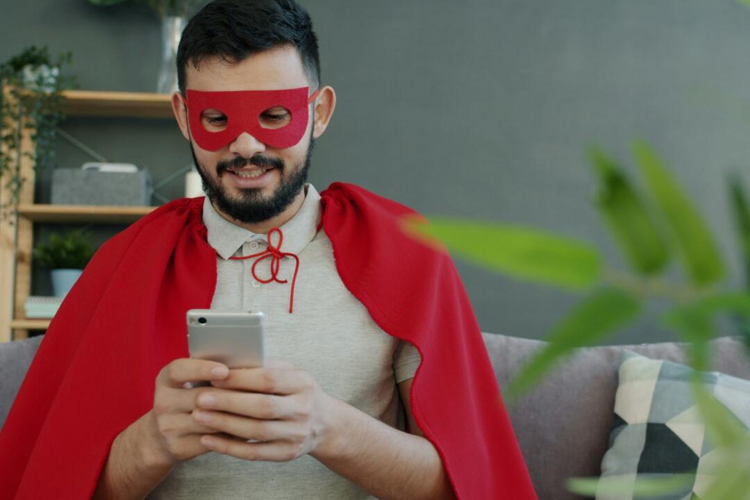 Excited man in red cape and mask is using smartphone touching screen enjoying good news online celebrating success. People and communication concept.