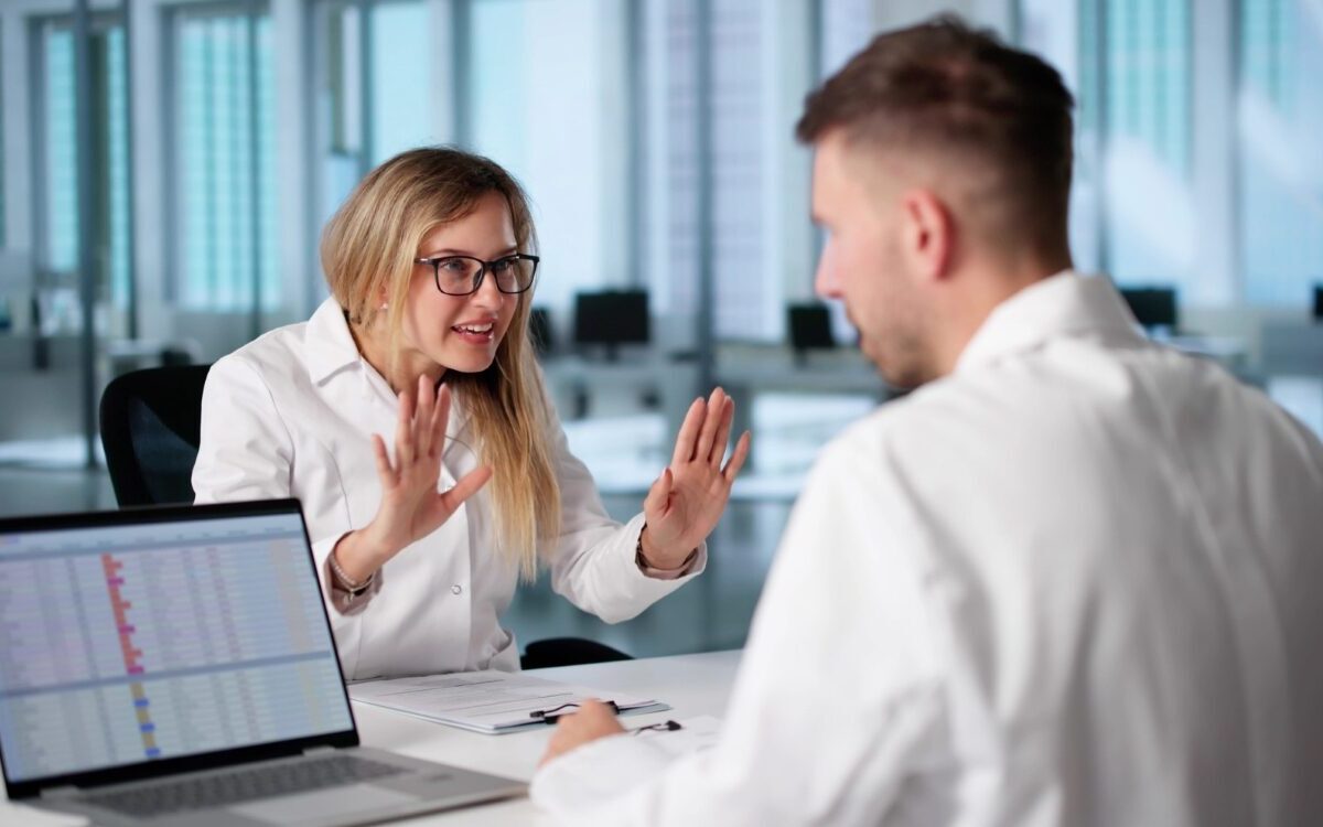 Two life science professionals in a laboratory office discussing data at a computer, illustrating tension in a research partnership and business decision-making.
