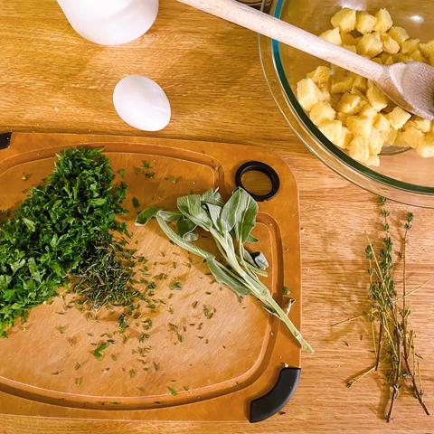 A chopping board with minced parsley, not yet minced sage, and thyme, an egg, a bowlful of yellow potato bread cubes and a canister of kosher salt