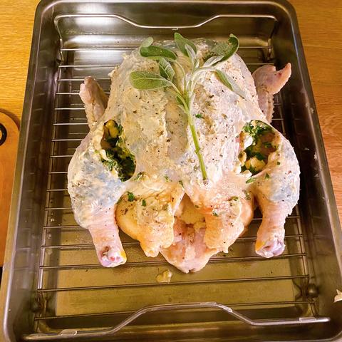 A raw chicken on a rack, in a steel roasting pan. The chicken skin has been cut open at the leg joints so I could insert stuffing under the skin of the breast and the legs. The whole chicken has been rubbed with butter and pepper, and there's a branch of sage spread out on the breast
