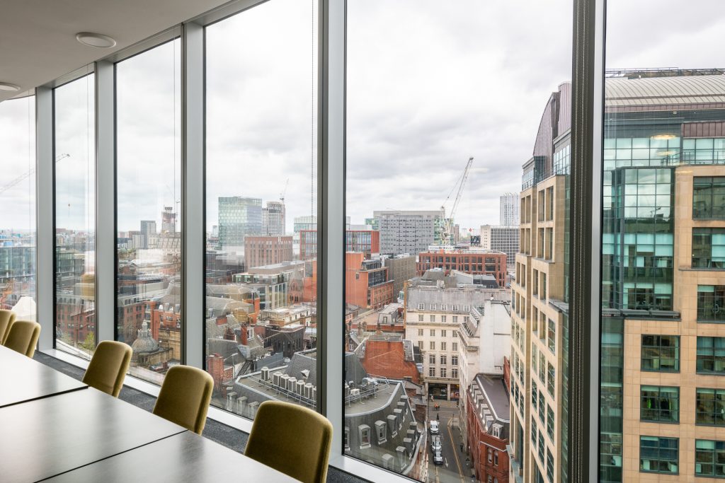 A meeting room with empty chairs around a table and behind them, floor to ceiling windows providing views of central Manchester. 