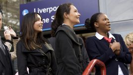 Three women in air hostess outfits outside of Liverpool Lime Street station gaze up at something off screen. The atmosphere is inspired