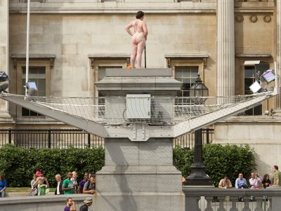 A curvy, naked woman stood on the Fourth Plinth. She has gold leaves on her breasts and holds a leaf over her crotch. People below wave and smile