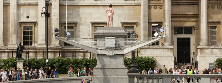 A curvy, naked woman stood on the Fourth Plinth. She has gold leaves on her breasts and holds a leaf over her crotch. People below wave and smile