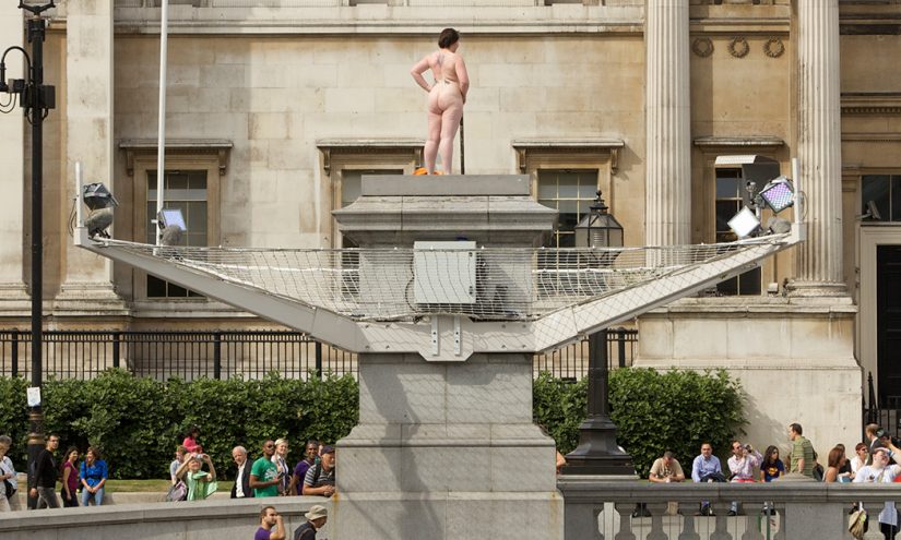 A curvy, naked woman stood on the Fourth Plinth. She has gold leaves on her breasts and holds a leaf over her crotch. People below wave and smile