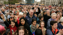 A dense crowd of spectators of all ages, packed into a city square lined with grand stone buildings, cheers and applauds with raised hands and excited expressions.