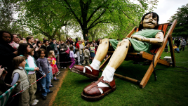 A giant wooden puppet of a young girl, dressed in a green outfit with white socks and brown sandals, reclines asleep in an oversized deckchair on grass beneath large trees. A crowd of onlookers, including several children, watches from behind a barrier tape line.