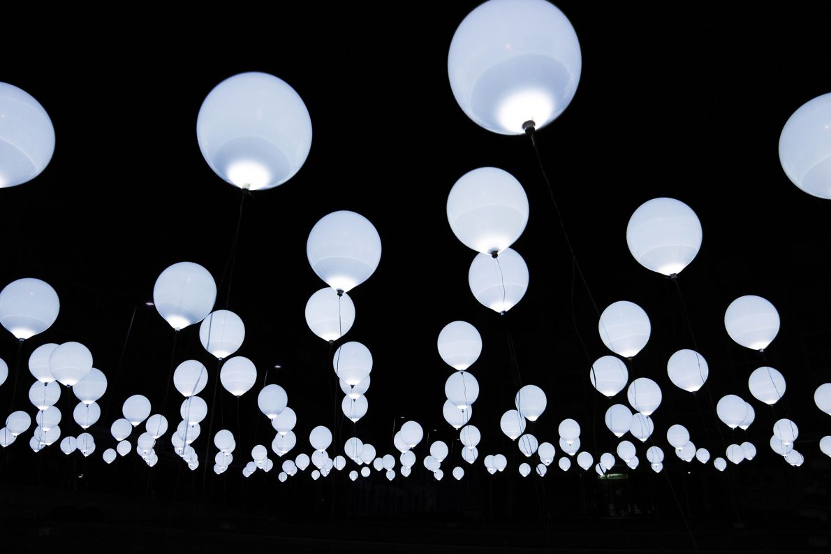 A multitude of glowing white balloons float against a dark night sky, creating a serene and ethereal atmosphere with their soft illumination.