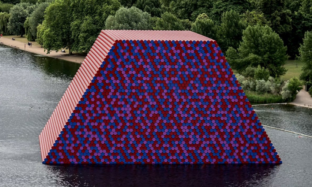 A large, colourful pyramid-shaped installation floats on a lake, composed of stacked red, blue, and purple barrels. Lush green trees line the shore.