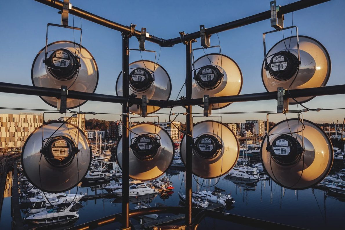 Nine round industrial lights are mounted on a metal frame overlooking a marina filled with docked boats. The sunset casts a warm glow over the scene.