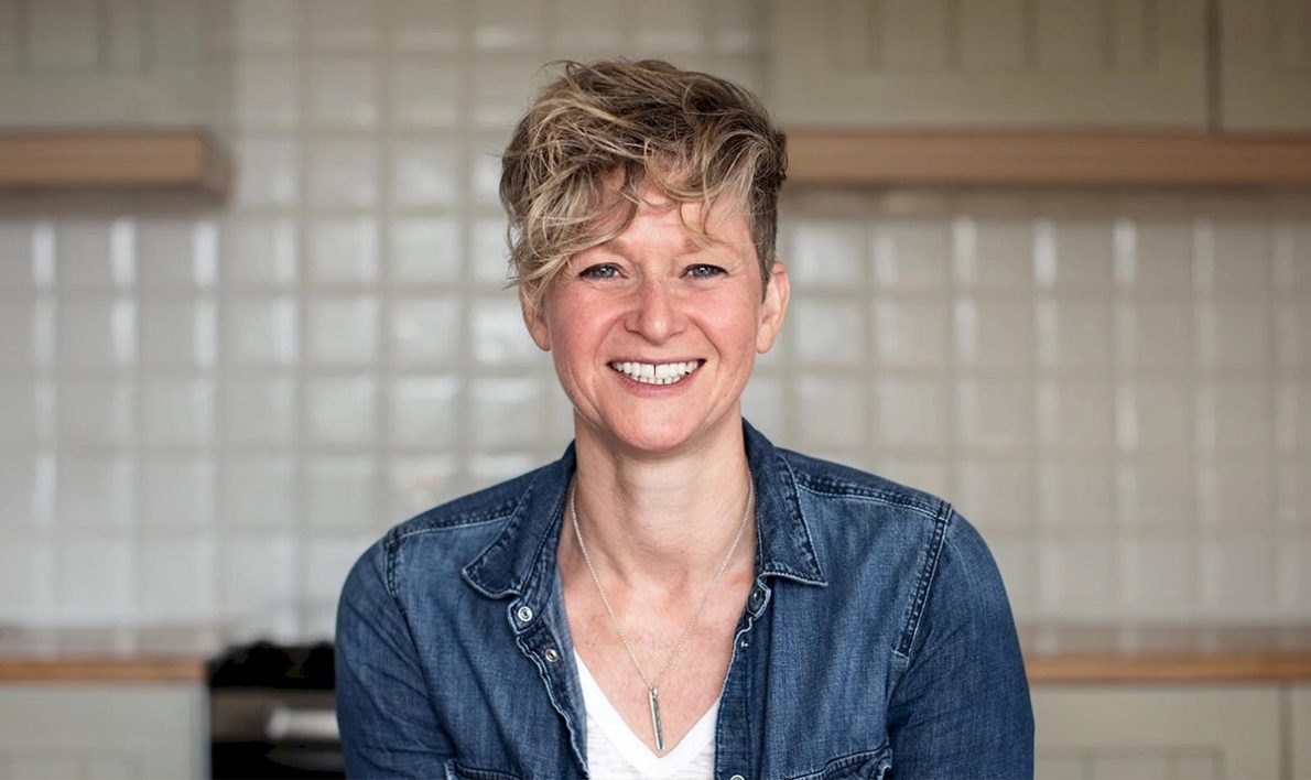 A white woman with a blonde pixie cut smiles at the camera. She is smiling and wearing a blue denim shirt with a white tank top.