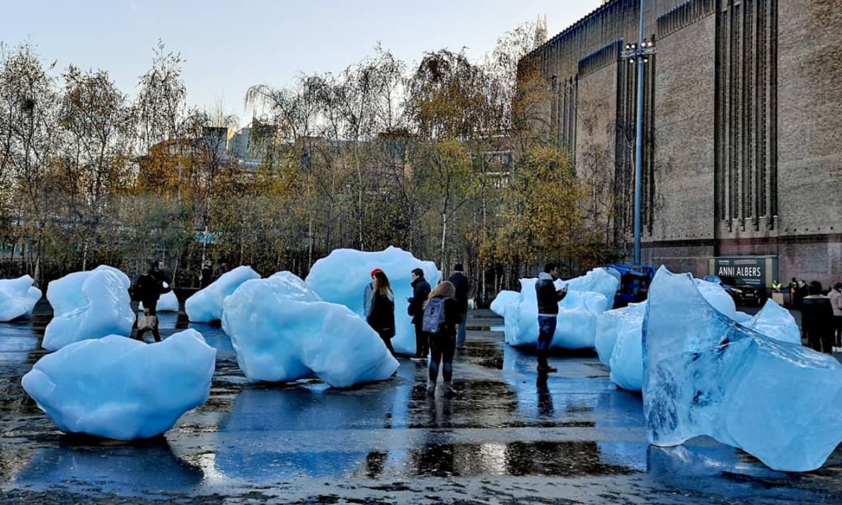 Large ice blocks scattered on a wet plaza, surrounded by people exploring the scene. Background features trees and the Tate Modern
