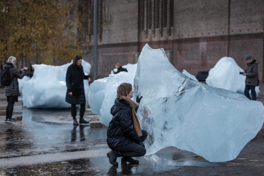 People interact with large ice blocks displayed on a wet city street. The scene conveys a sense of curiosity and environmental awareness. The focus of the image shows a woman crouched in front of a large chunk of ice, she has her hand on the ice.