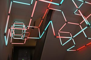 Suspended geometric neon installation with glowing blue and red tubes in a dark hallway. The arrangement creates a modern, artistic ambiance. It resembles Geomag - the classic building toy with coloured magnetic sticks and little metal balls