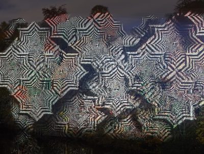 An intricate pattern of overlapping geometric shapes in black and white is projected onto a dark forest backdrop, creating a mesmerising, hypnotic effect.