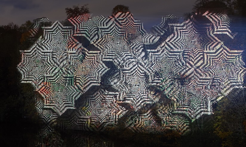 An intricate pattern of overlapping geometric shapes in black and white is projected onto a dark forest backdrop, creating a mesmerising, hypnotic effect.